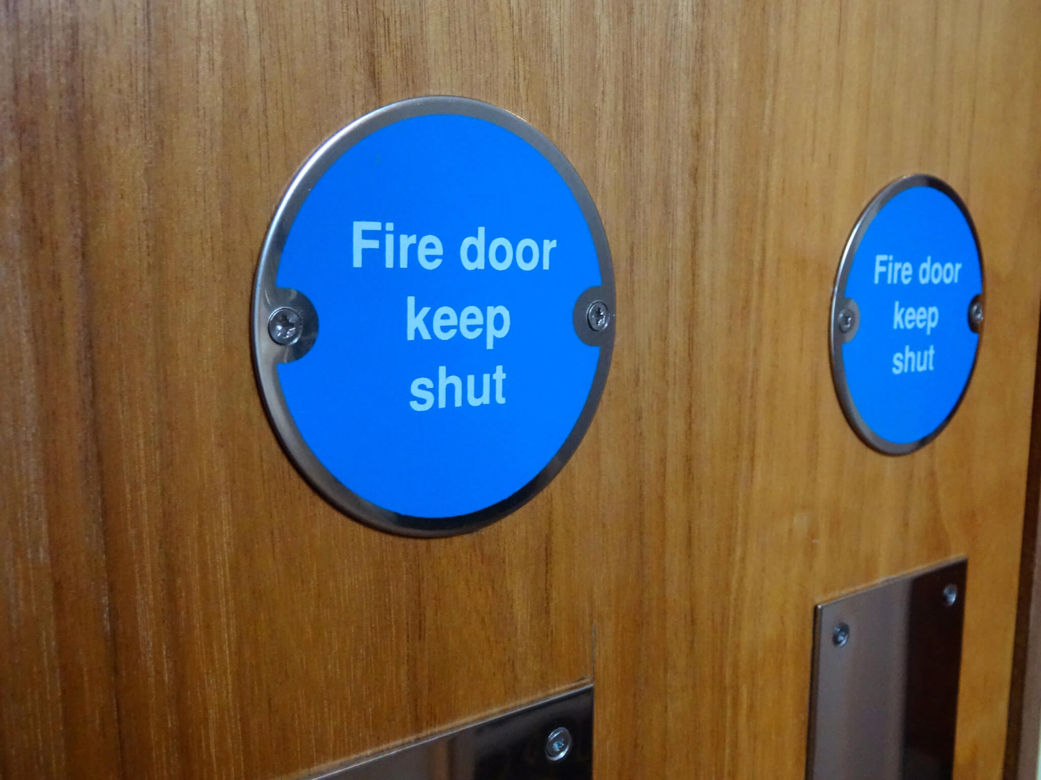 Photo showing a circular blue sign with white writing saying 'Fire Door Keep Shut '.  This safety sign has been screwed onto a wooden fire door.